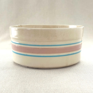 May include: Cream-colored ceramic bowl with horizontal stripes. The bowl features a wide pink stripe bordered by two thin blue stripes. The stripes encircle the bowl. The bowl has a simple, rounded shape and a smooth, glossy finish.