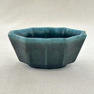 May include: A teal ceramic bowl with a geometric, multi-sided design. The bowl has a glossy finish and a slightly textured surface. The sides are vertically ribbed, adding visual interest. Likely for serving or display, it's a stylish piece.