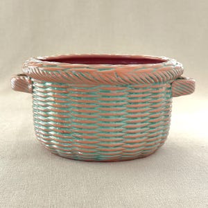Puede incluir: Una cesta de cerámica con diseño tejido, con exterior coral y interior turquesa. El borde y las asas tienen una textura de cuerda. La cesta está diseñada para uso decorativo o funcional.