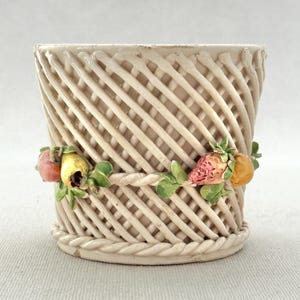 Puede incluir: Cesta de cerámica color crema con diseño tejido. La cesta está adornada con una guirnalda de frutas coloridas, incluyendo fresas, peras y naranjas. La base y el borde tienen una textura de cuerda.