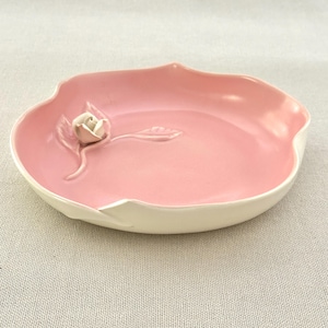 May include: A pink ceramic dish with a white rim and a raised rose design. The dish has a smooth, glossy finish and a unique, organic shape. The rose is a delicate, three-dimensional accent.