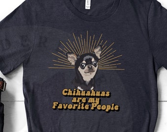May include: A heather navy blue t-shirt with a graphic of a chihuahua and the text "Chihuahuas are my Favorite People". The shirt is laid out on a white surface with a brown leather bag, black boots, sunglasses, and brown pants.