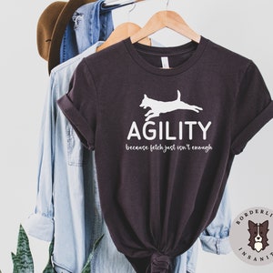 Może przedstawiać: Szary t-shirt z białym siluetem psa skaczącego przez przeszkodę. Pod psem znajduje się napis "Agility", a pod nim napis "because fetch just isn't enough". T-shirt wisi na drewnianym wieszaku.