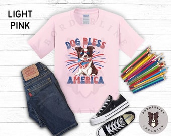 May include: A light pink t-shirt with a graphic of a dog wearing sunglasses and a bandana with the text "Dog Bless America" and fireworks in the background.