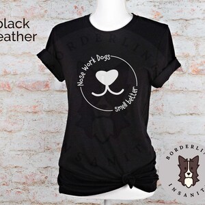Może przedstawiać: Czarny t-shirt z melanżu z białym nadrukiem nosa psa i tekstem "Nose Work Dogs Smell Better". T-shirt ma okrągły dekolt i kr&oacute;tkie rękawy. Nadruk znajduje się w k&oacute;łku z białym obramowaniem.