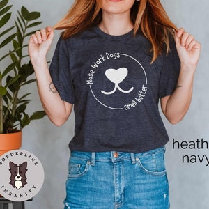 Może przedstawiać: Koszulka w kolorze granatowym z odcieniem heathered z białym nadrukiem nosa psa i tekstem "Nose Work Dogs - Smell Better".