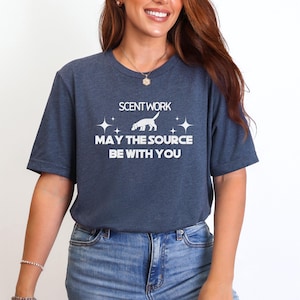 May include: A heather navy blue short-sleeve t-shirt featuring white text that reads SCENT WORK MAY THE SOURCE BE WITH YOU, accompanied by a white graphic of a dog and four-pointed star accents centered on the chest area of the garment.
