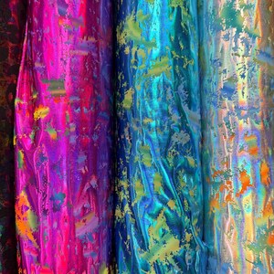 Puede incluir: Primer plano de varios rollos de tela brillante en colores vibrantes. La tela tiene un brillo met&aacute;lico y est&aacute; pintada con pinceladas abstractas en tonos de rosa, azul, verde y dorado. Los rollos est&aacute;n muy juntos, mostrando la textura y las variaciones de color.