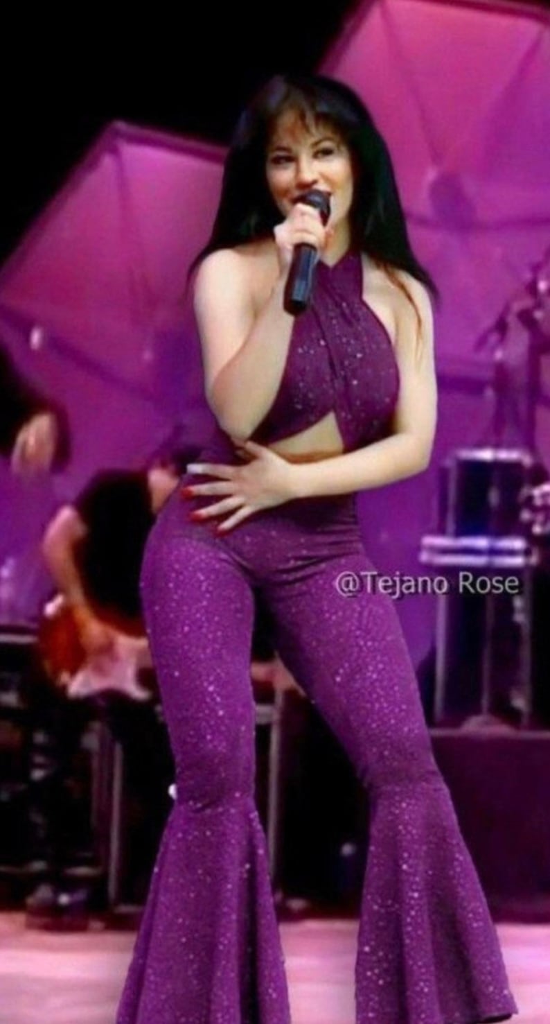 May include: A woman in a sparkly purple jumpsuit with a halter top sings into a microphone on stage. The background is purple and there is a drum set behind her.