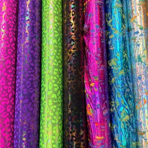 Puede incluir: Una colecci&oacute;n de rollos de tela en varios colores, incluyendo rosa, morado, verde, negro, azul y plateado. La tela tiene un acabado brillante y met&aacute;lico con un estampado de leopardo. Los rollos est&aacute;n dispuestos verticalmente, mostrando los diferentes colores y texturas.