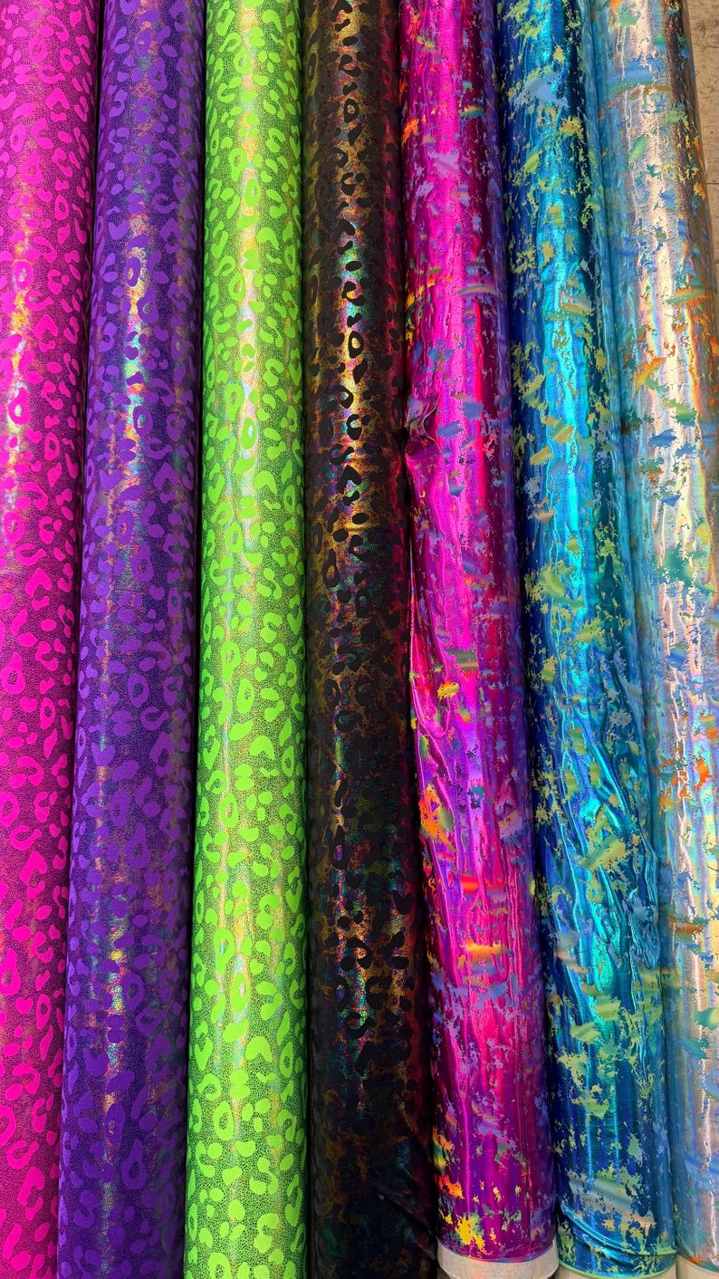 Puede incluir: Una colecci&oacute;n de rollos de tela en varios colores, incluyendo rosa, morado, verde, negro, azul y plateado. La tela tiene un acabado brillante y met&aacute;lico con un estampado de leopardo. Los rollos est&aacute;n dispuestos verticalmente, mostrando los diferentes colores y texturas.