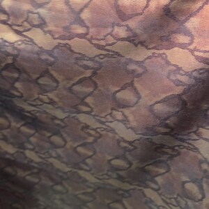 Op de afbeelding: Close-up van een stof met een herhalend patroon van donkerbruine, ovale vormen omlijnd in een donkerdere tint, tegen een gevlekt achtergrond van bruin- en bruintinten. De stof heeft een getextureerd uiterlijk.