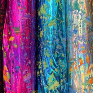 Puede incluir: Primer plano de varios rollos de tela iridiscente en colores vibrantes. La tela tiene un brillo met&aacute;lico con salpicaduras de color, incluyendo rosa, azul, verde y dorado. El material parece texturizado y adecuado para varios proyectos de artesan&iacute;a.
