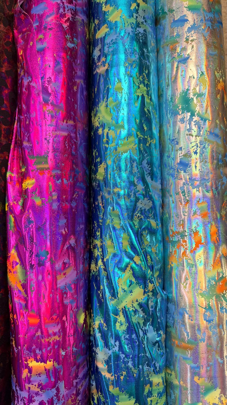 Puede incluir: Primer plano de varios rollos de tela iridiscente en colores vibrantes. La tela tiene un brillo met&aacute;lico con salpicaduras de color, incluyendo rosa, azul, verde y dorado. El material parece texturizado y adecuado para varios proyectos de artesan&iacute;a.