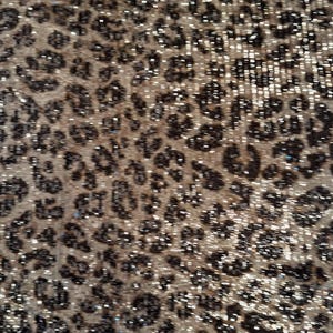 May include: Close-up of a fabric with a leopard print pattern. The design features dark brown spots on a tan background, with a shimmering, glittery effect throughout the fabric.