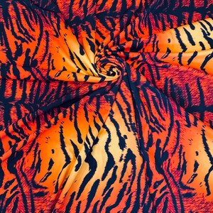 Puede incluir: Primer plano de una tela con un estampado de rayas de tigre. La tela presenta una mezcla de colores naranja, rojo y negro, creando un dise&ntilde;o llamativo. El material est&aacute; recogido en el centro, mostrando su textura y ca&iacute;da.