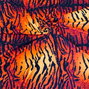 Puede incluir: Tela vibrante con un estampado de rayas de tigre. El dise&ntilde;o presenta gruesas rayas negras sobre un fondo naranja brillante y amarillo. La tela est&aacute; plegada en el centro, creando un efecto arremolinado.