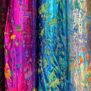 Puede incluir: Primer plano de varios rollos de tela iridiscente en colores vibrantes. La tela tiene un brillo met&aacute;lico y est&aacute; salpicada de varios colores, incluyendo rosa, azul y dorado. La tela se utiliza probablemente para ropa o manualidades.