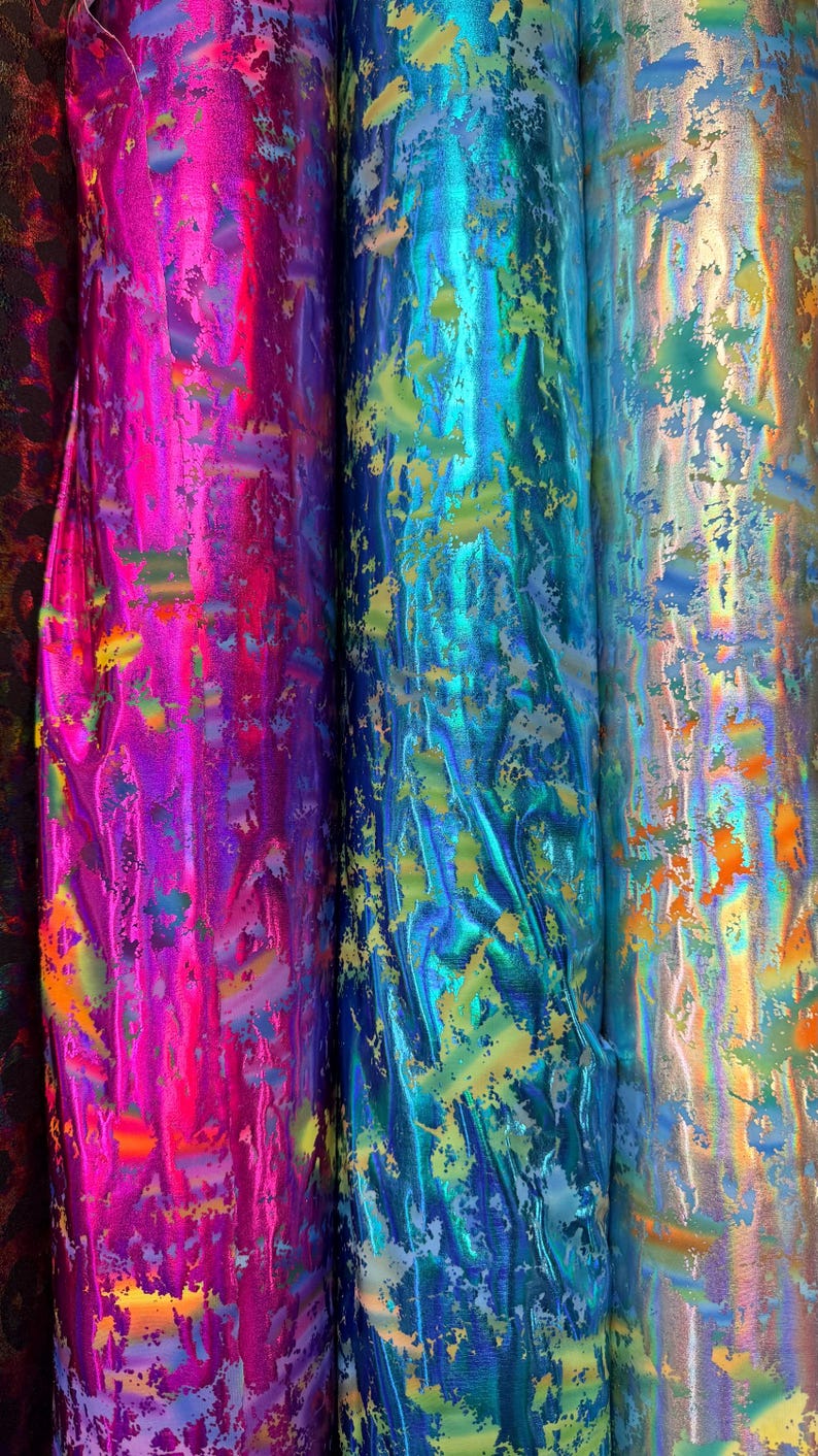 Puede incluir: Primer plano de varios rollos de tela iridiscente en colores vibrantes. La tela tiene un brillo met&aacute;lico y est&aacute; salpicada de varios colores, incluyendo rosa, azul y dorado. La tela se utiliza probablemente para ropa o manualidades.