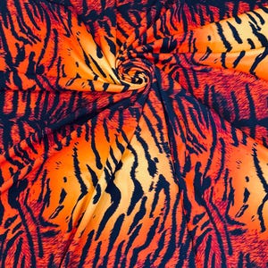 Puede incluir: Primer plano de una tela con un estampado de rayas de tigre. La tela presenta audaces rayas naranjas y negras, creando un efecto visual llamativo. El material est&aacute; recogido en un remolino, mostrando el flujo del patr&oacute;n.