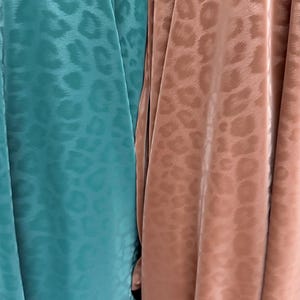 May include: Two pieces of fabric hanging side by side. The left piece is a vibrant teal colour, and the right piece is a muted dusty rose colour. Both fabrics feature a tonal, repeating leopard print pattern with a smooth, satin-like finish.