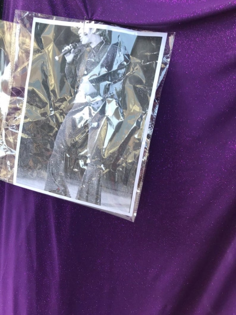 May include: A close-up of a purple sparkly fabric with a black and white photo of a person singing into a microphone. The photo is in a clear plastic sleeve.