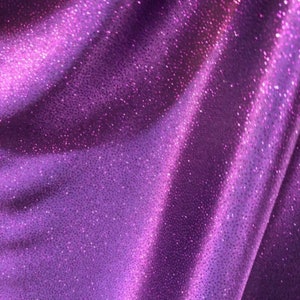 May include: Purple fabric with a sparkly, glittery texture. The fabric is smooth and has a slight sheen.