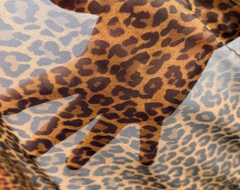 May include: Close-up of a sheer fabric with a leopard print pattern. The fabric is a light brown color with dark brown spots. The fabric is held up, and the hand is visible through the material.