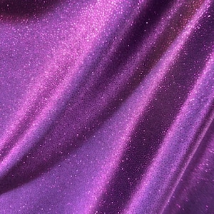 May include: Purple fabric with a sparkly, glittery texture. The fabric is folded and draped, showing the shimmery effect of the material.
