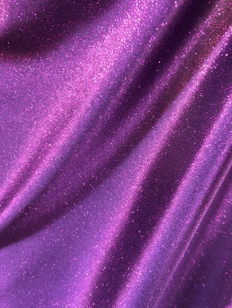 May include: Purple fabric with a sparkly, glittery texture. The fabric is folded and draped, showing the shimmery effect of the material.