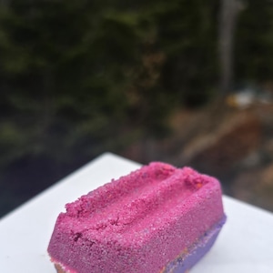 May include: A rectangular bath bomb with a layered design. The top is a textured, vibrant pink, with a middle layer of orange, and a purple base. The bath bomb sits on a white surface.