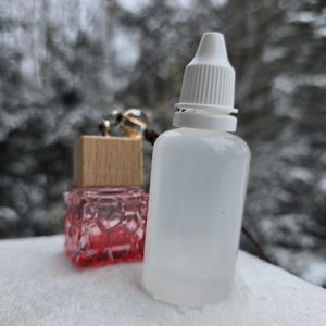 May include: A clear glass bottle with a white dropper top and a red glass bottle with a wooden top. Both bottles are sitting on a white surface.
