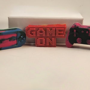 May include: Three colorful, hand-crafted, wax game controllers. The controllers are shaped like a handheld game console, a game controller, and a sign that says "GAME ON".