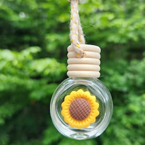 May include: A clear glass bottle with a sunflower design hanging from a beige rope with wooden beads. The bottle is filled with a clear liquid and has a wooden lid.