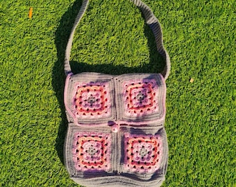 Puede incluir: Un bolso de hombro de crochet gris con detalles en rosa y blanco. El bolso está hecho de cuadrados con un patrón floral.