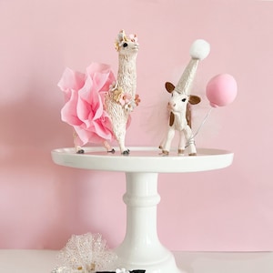 May include: Three figurines of a llama, a cow, and a calf, all dressed in party attire. The llama is wearing a pink tutu and a flower crown. The cow is wearing a white party hat and a tulle skirt. The calf is wearing a white tulle skirt and a flower crown. The figurines are on a white cake stand.