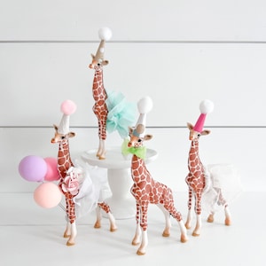 May include: Four brown and white giraffe figurines wearing colourful party hats and tulle skirts. The giraffes are arranged on a white surface with pink, purple and white balloons.