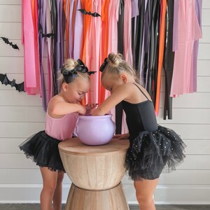 May include: Two girls dressed in black and pink tutus are standing in front of a wall decorated with colourful streamers and bats. They are holding a purple cauldron and looking at it.