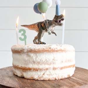 May include: A round cake with white frosting, decorated with a toy dinosaur, balloons, and candles. The candles spell out the number 3. The cake sits on a wooden cake stand, perfect for a birthday celebration.