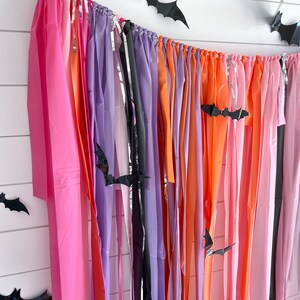 May include: A Halloween party backdrop with a colourful streamer garland made of pink, orange, purple, and black tissue paper. Black paper bats are strung throughout the garland.