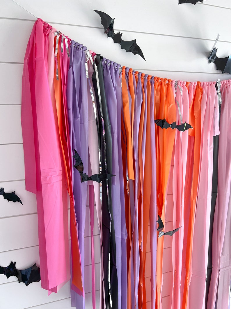 May include: A Halloween party backdrop with a colourful streamer garland made of pink, orange, purple, and black tissue paper. Black paper bats are strung throughout the garland.