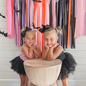 May include: Two young girls wearing black tulle tutus with gold glitter, pink and black tank tops, and black bows in their hair. They are posing in front of a colourful streamer backdrop with bats.