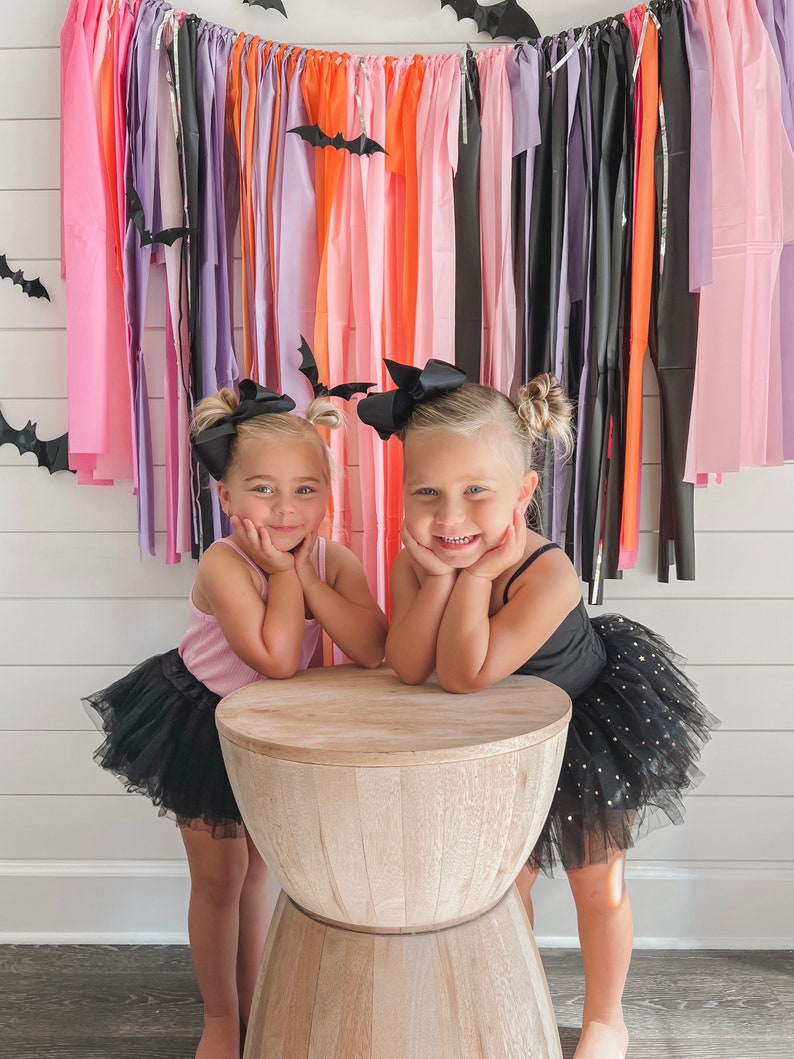 May include: Two young girls wearing black tulle tutus with gold glitter, pink and black tank tops, and black bows in their hair. They are posing in front of a colourful streamer backdrop with bats.