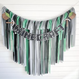 May include: A Minecraft themed party banner with the word "MINECRAFT" in black letters with a pixelated design. The banner is strung with green, gray, and black tissue paper streamers.