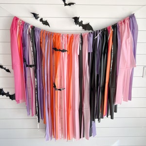 May include: A Halloween party backdrop with a streamer garland made of pink, orange, purple, and black tissue paper. Black paper bats are hanging from the white wall behind the garland.