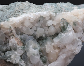 May include: Close-up of a mineral specimen featuring a cluster of clear quartz crystals. The crystals are surrounded by a matrix of pale green and gray rock. Some crystals have a green hue.