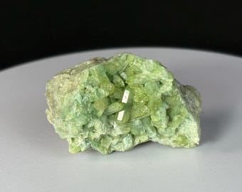 May include: A raw, uncut mineral specimen featuring a cluster of light green crystals. The crystals have a prismatic shape and are clustered together, creating a textured surface. The mineral has a pale green hue.