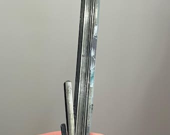 May include: A slender, elongated mineral crystal with a dark, metallic gray color and a vertically striated, reflective surface. The crystal features a sharp, pointed tip and a smaller, secondary splintered segment attached near the base.