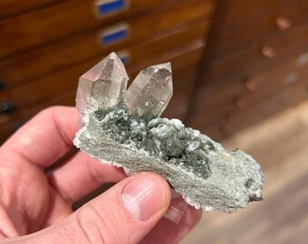 May include: A hand holding a cluster of clear quartz crystals with a green mineral base. The crystals are pointed and translucent, with a light green hue. The base is a rough, textured, gray-green color.