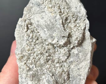 May include: A light gray rock formation with small, crystalline structures clustered across its surface. The rock has a rough, textured appearance, with variations in color and tone. The overall shape is irregular, with a jagged edge.